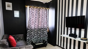 A living room with a black and white striped wall, a flat-screen TV, a sofa with red cushions, and a patterned curtain. - Good Deal 2Br At 16Th Floor Suites @Metro Apartment (Bandung)