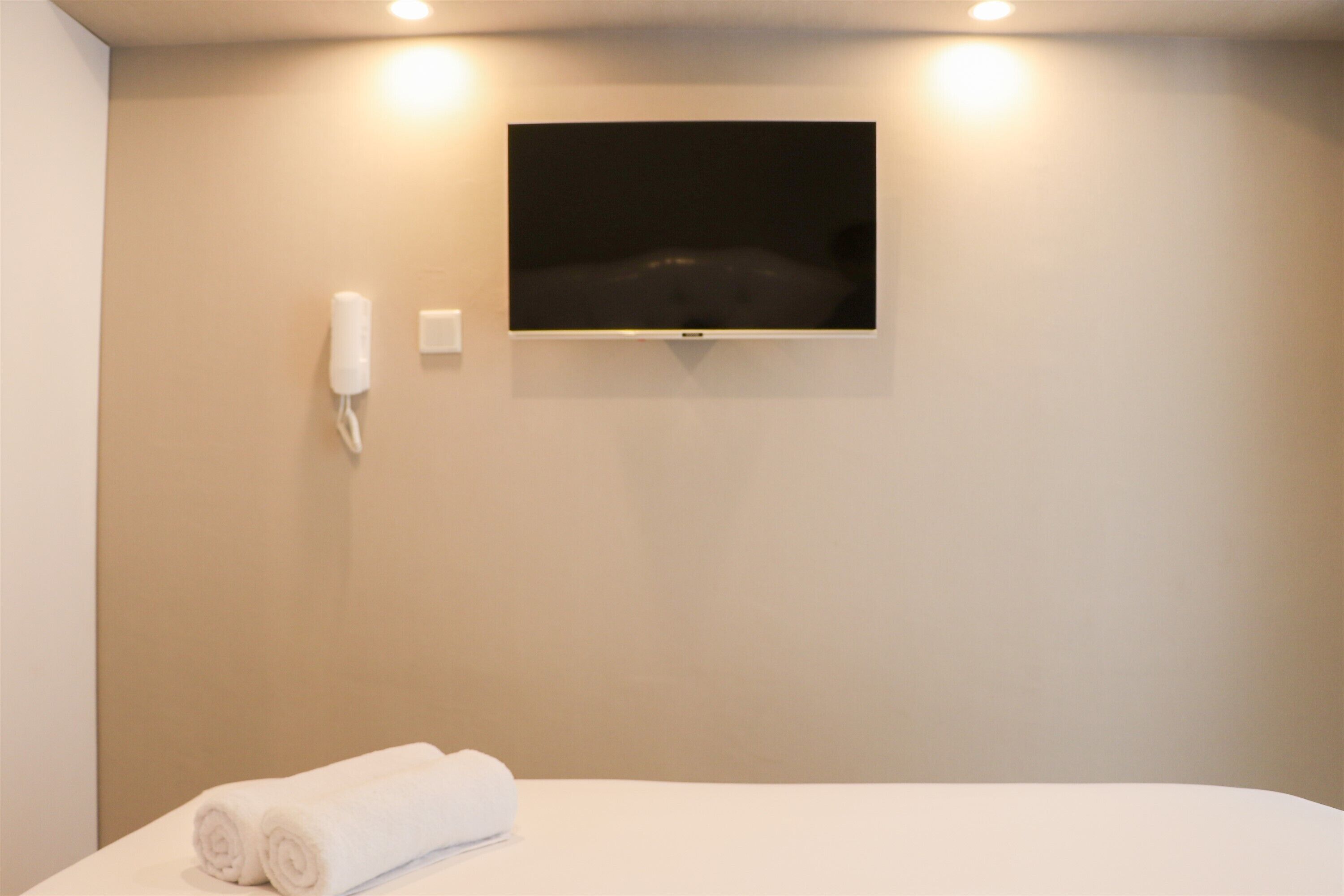 A hotel room with a bed, a wall-mounted TV, a telephone, and a rolled-up towel on the bed.