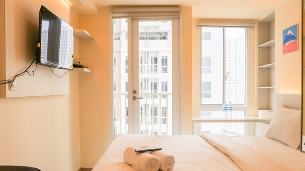 A hotel room with a bed, a TV mounted on the wall, a window with a view of buildings, and a shelf with bottles. - Nice And Homey Studio Tokyo Riverside Pik 2 Apartment (Teluknaga)