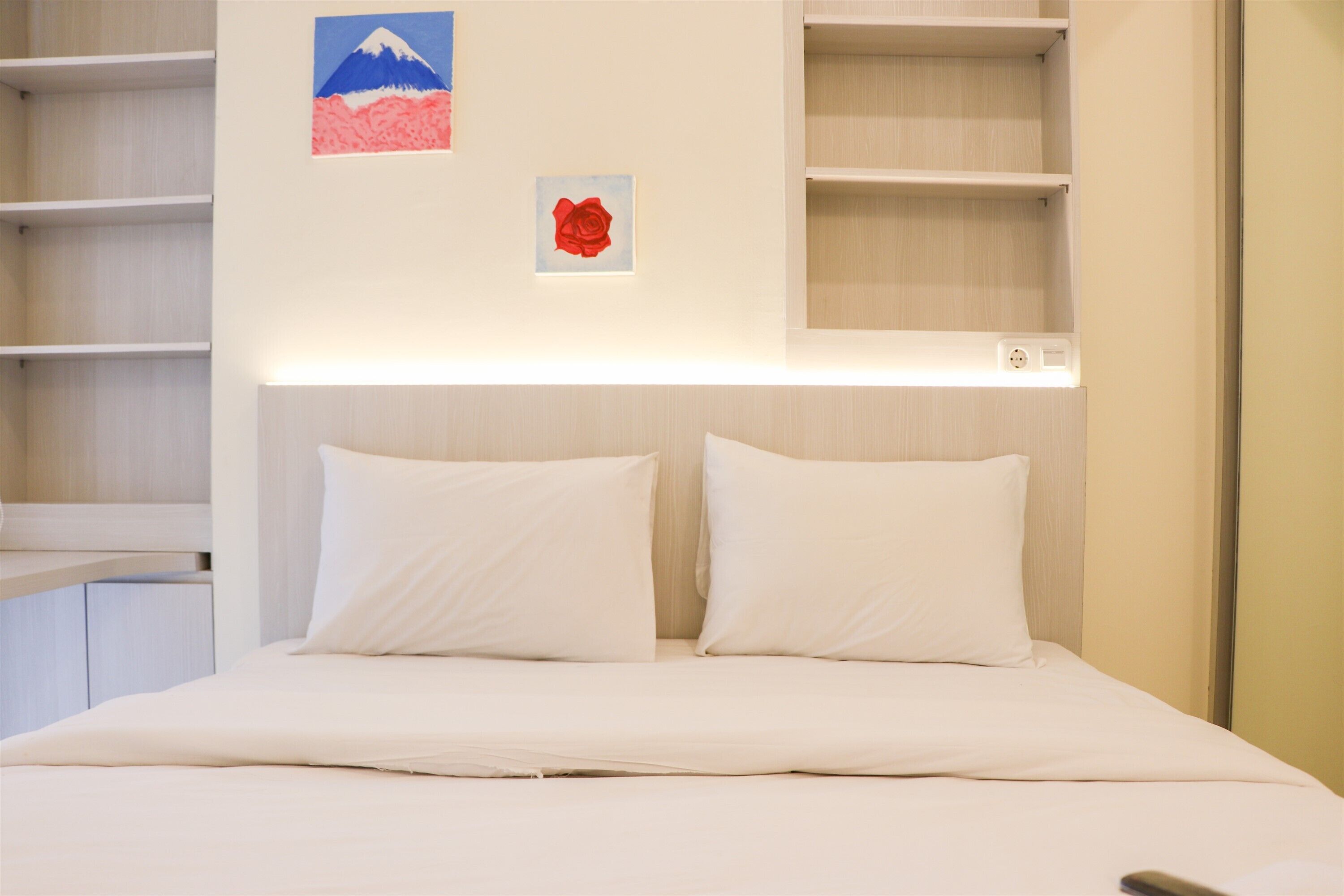 A hotel room with a bed, white pillows, a mounted shelf with a power outlet, and two wall-mounted artworks (a mountain and a rose).