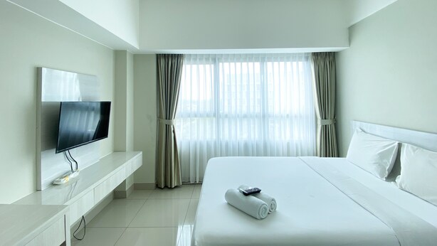 A hotel room with a bed, a TV on a stand, a window with curtains, and a white wall. - Restful And Simply Studio Springlake Summarecon Bekasi Apartment (Bekasi)