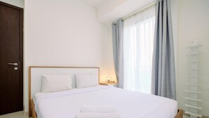 A neatly made bed with white bedding and a folded towel, a wooden headboard, a bedside table with a lamp, and a window with grey curtains.