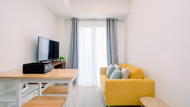 A modern living room with a yellow sofa, a TV on a wooden table, and a view through sheer curtains.