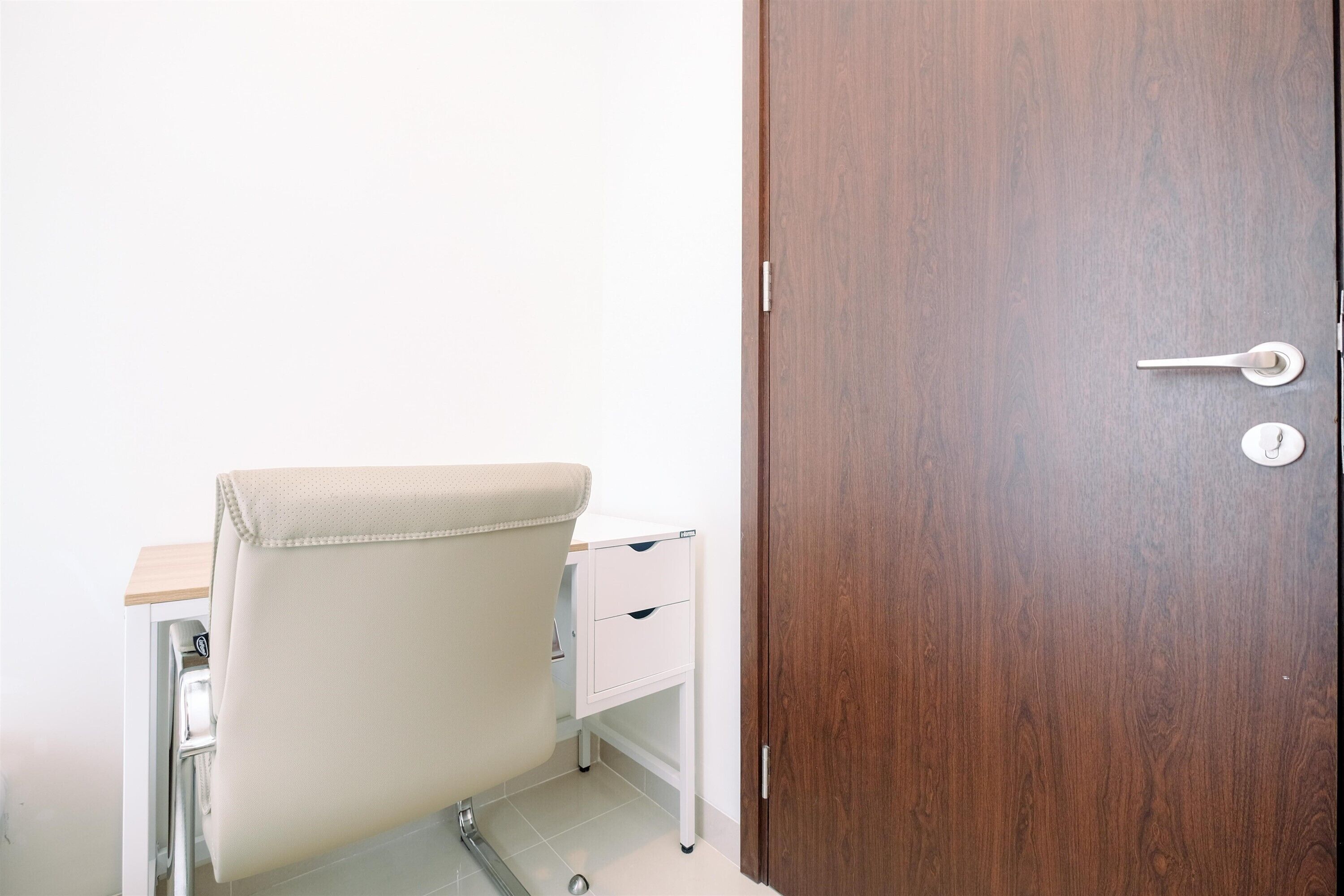 A room with a wooden door, a white desk with drawers, and a chair with a beige cushion.
