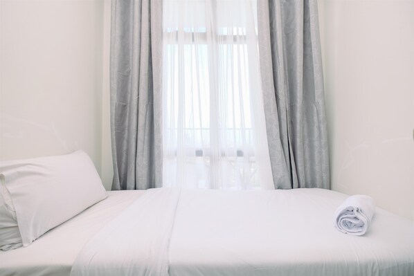 A neatly made bed with white linens and a rolled towel, a window with sheer curtains, and a plain wall.