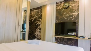 A hotel room with a bed, a television, a clock, and a desk with a computer. - Comfort And Modern Look 3Br Sky House Bsd Apartment (Pagedangan)