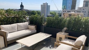 Una terraza en la azotea con un sofá, una mesita y una silla. La terraza ofrece vistas de un paisaje urbano con edificios y una estructura en forma de cúpula.