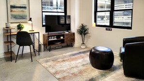 Una sala de estar moderna con un televisor de pantalla plana, un escritorio con una lámpara, una planta en maceta y un reposapiés de cuero.