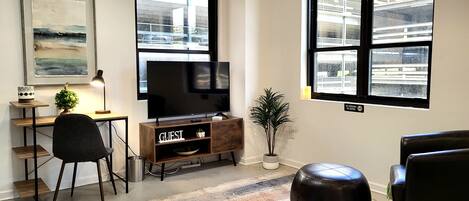 Un salon moderne avec un téléviseur à écran plat, un bureau avec une lampe, une plante en pot et un pouf en cuir.
