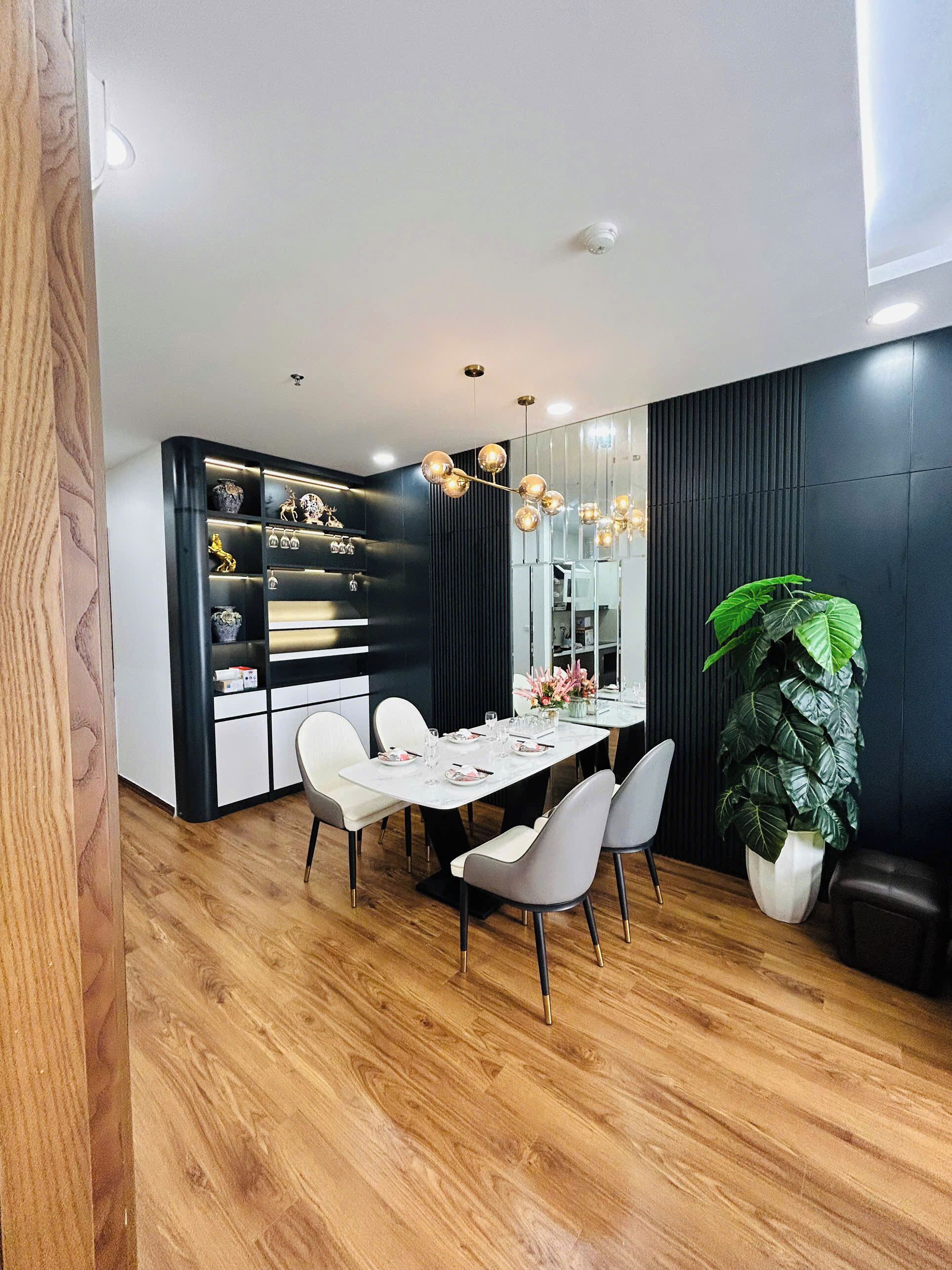 A modern dining area with a white table set for four, surrounded by grey chairs. A black shelving unit with decorative items is to the left, and a large mirror reflects the room. A green plant is in the corner.