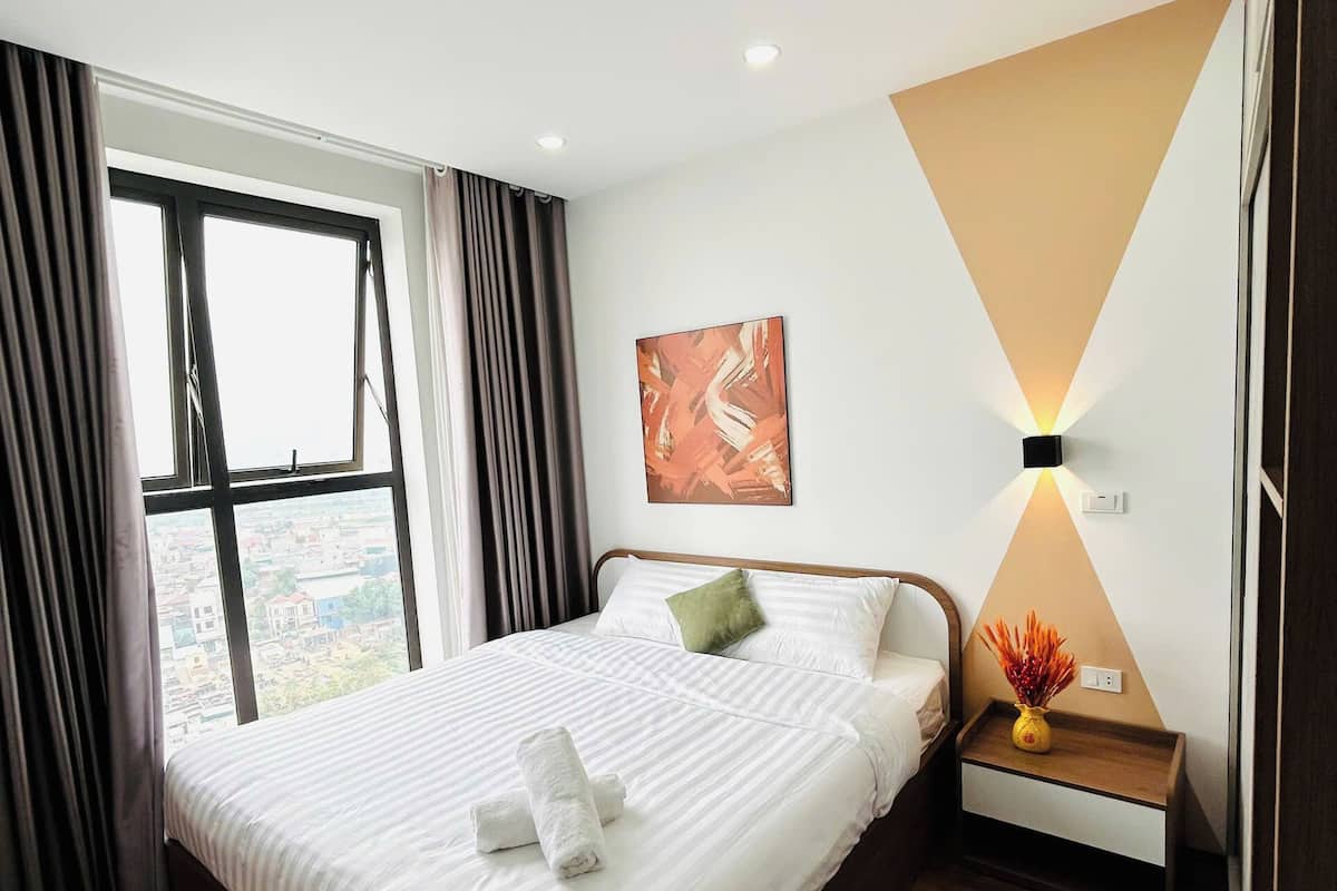 A modern hotel room with a large bed, a nightstand with a vase, and a view of the city through a window.