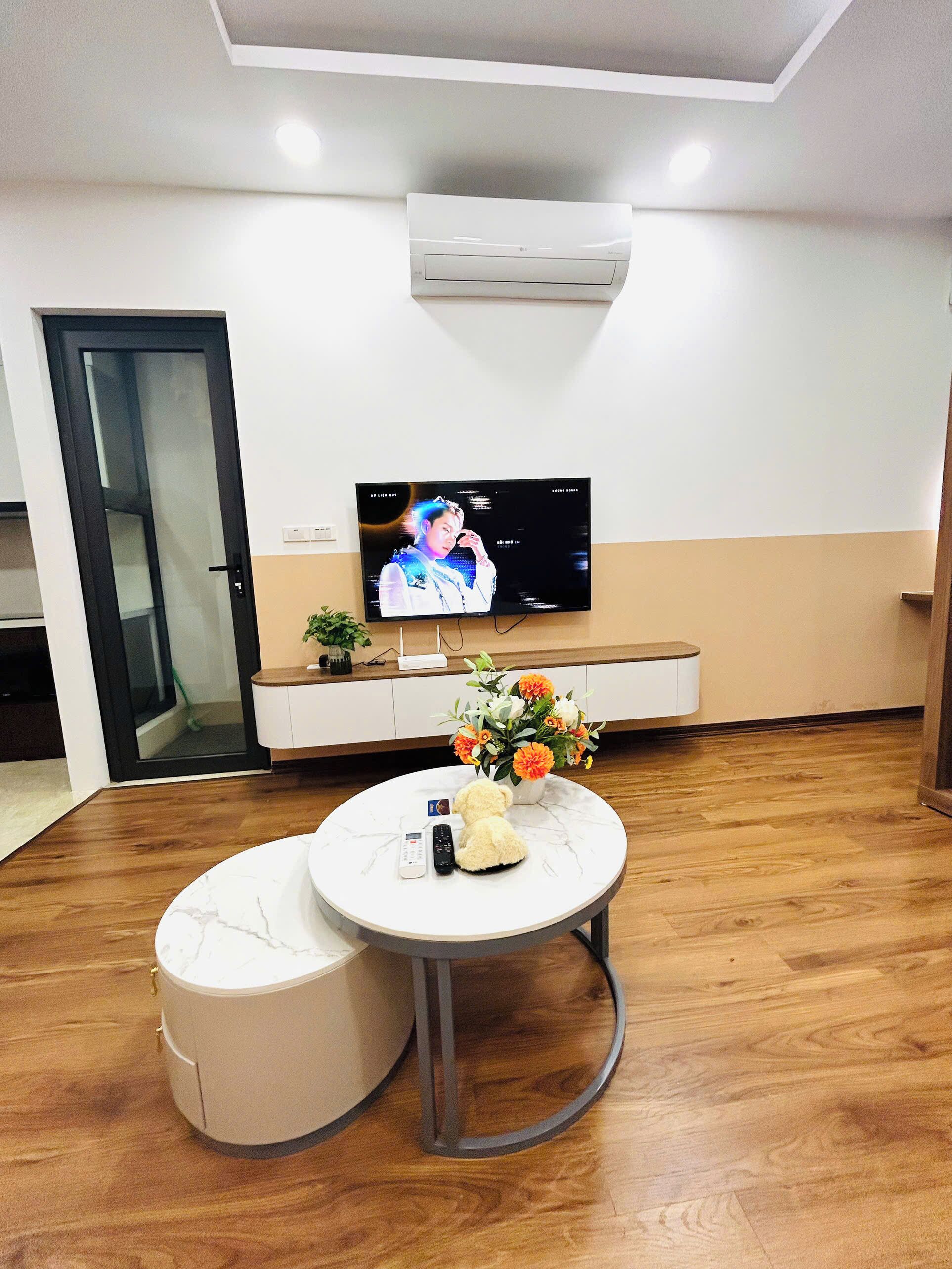 Un salon moderne avec un téléviseur à écran plat fixé à un mur blanc, une table basse ronde ornée d’un vase de fleurs, un canapé et un plancher en bois.
