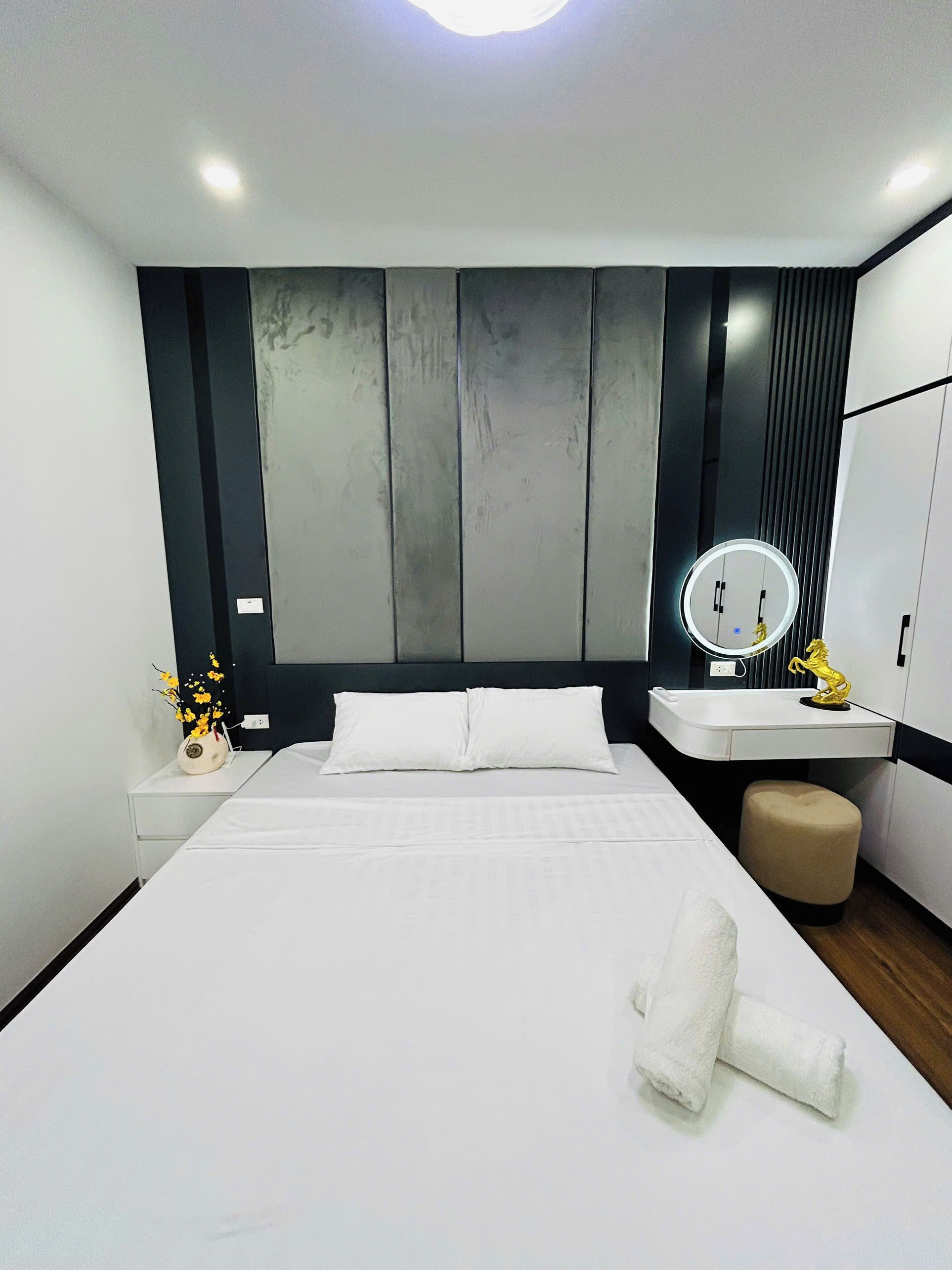 A modern hotel room with a large bed, a bedside table with a vase of flowers, a round mirror, and a small stool.