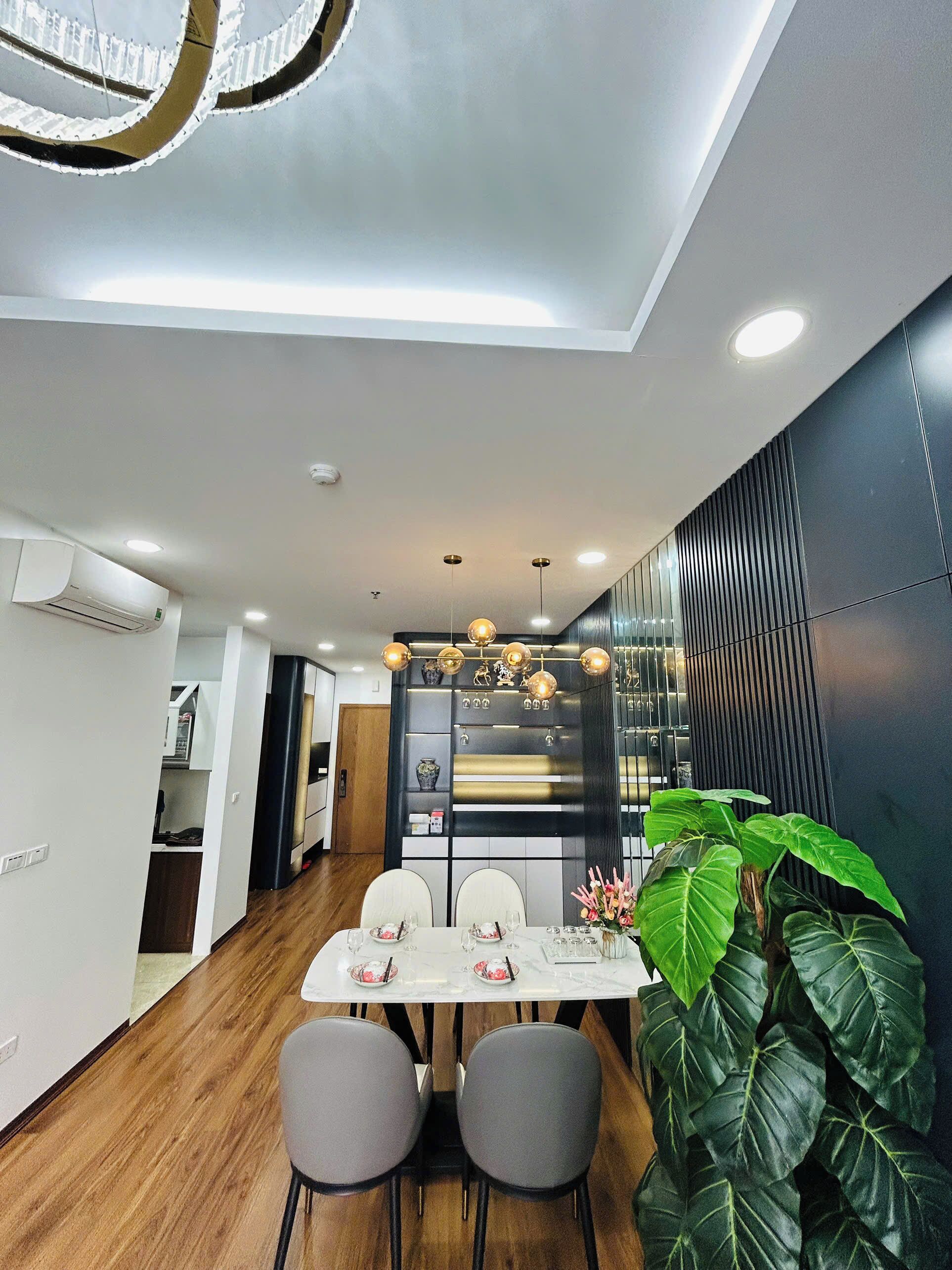 A modern dining area with a wooden floor, a white table, and four grey chairs. There is a large green plant on the right.