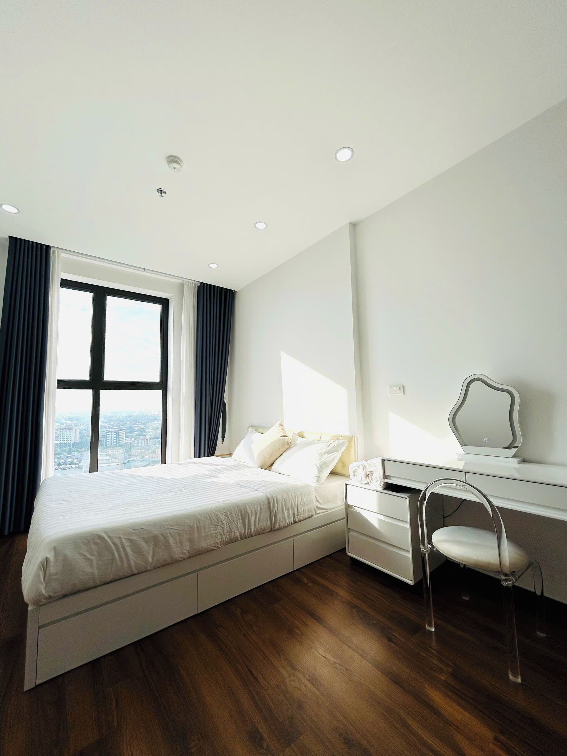 A modern hotel room with a large bed, a desk with a chair, and a view of the city through a large window.
