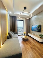 Una sala moderna con un sofá modular, una mesa redonda con flores, un televisor de pantalla plana montado en la pared y un balcón con vistas.