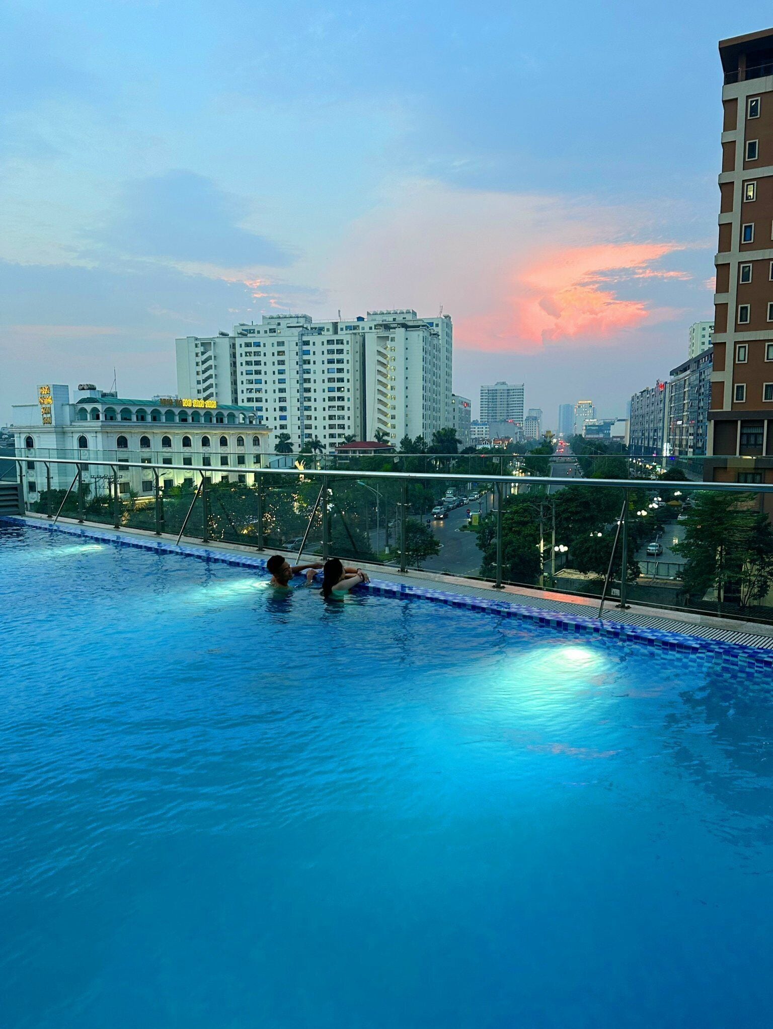 A swimming pool with two people in the water, surrounded by city buildings and a sunset sky.
