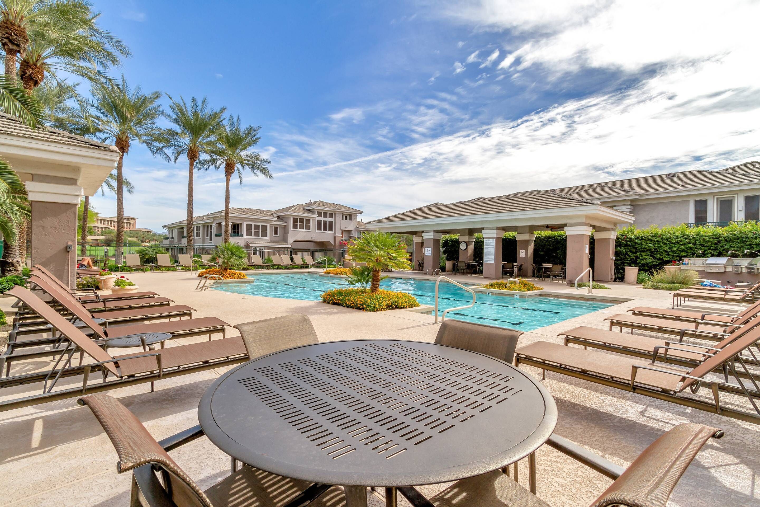 A poolside area with lounge chairs and a table, surrounded by palm trees and a clear sky.