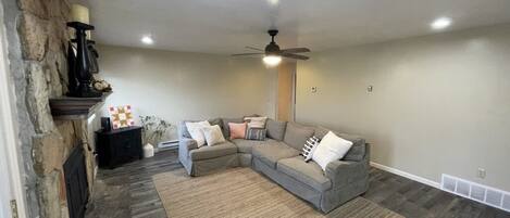 A living room with a sectional sofa, a fireplace, a ceiling fan, and a stone wall.