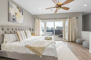 A bedroom with a large bed, a ceiling fan, a city view through a sliding door, and a modern abstract painting on the wall.