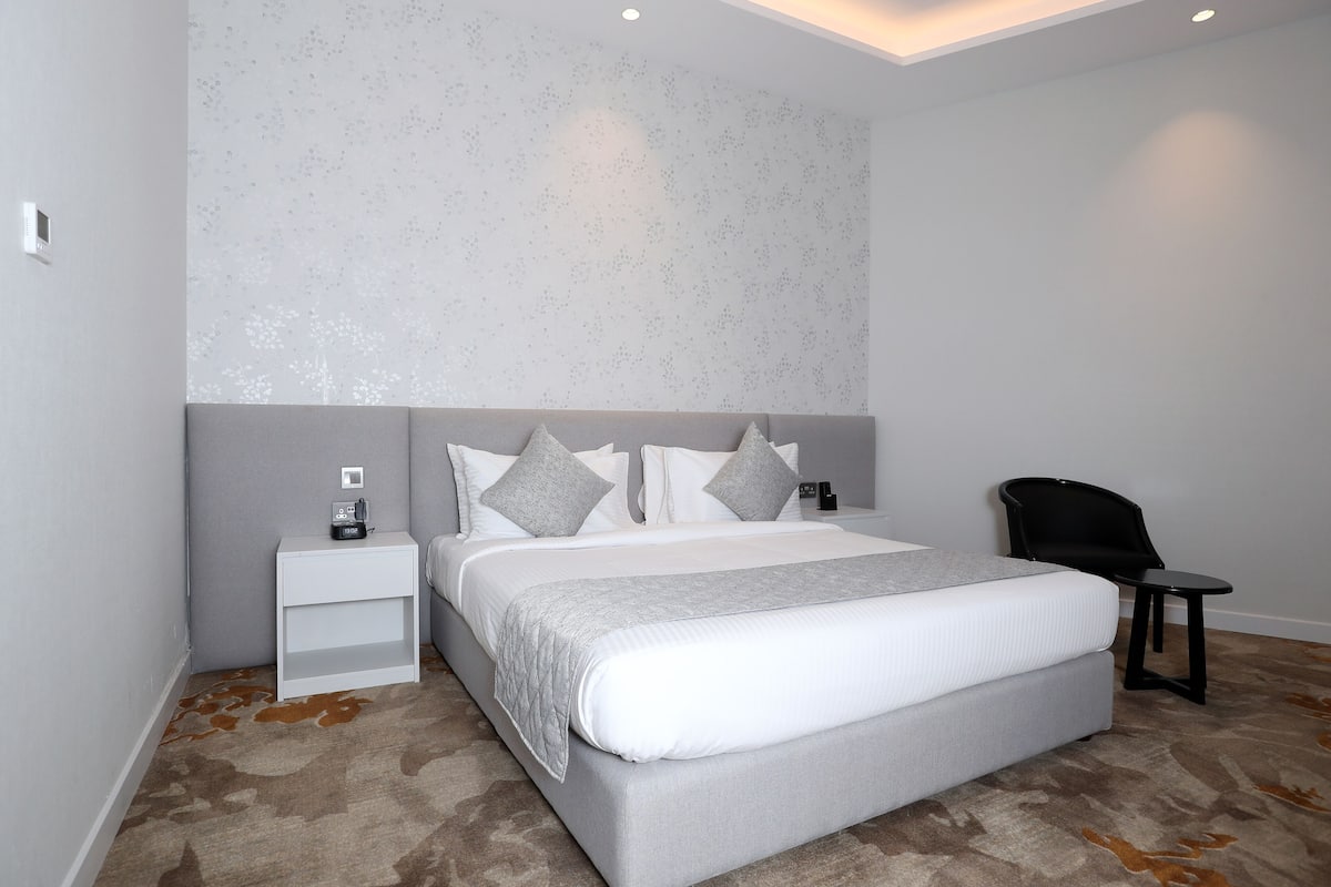 A modern bedroom with a large bed, grey and white bedding, a nightstand with a phone, and a small table.