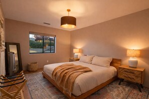 A bedroom with a large bed, two bedside tables with lamps, a hanging light fixture, a window with a view, a mirror, a heater, and a patterned rug.