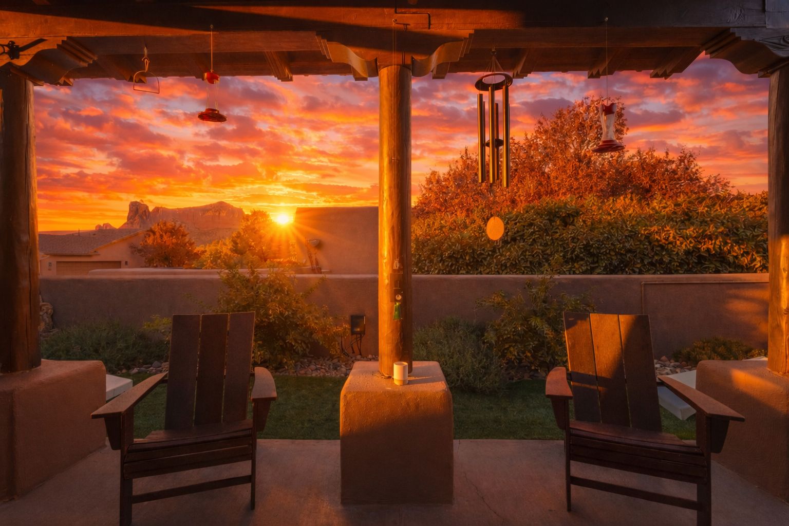 Una terrazza esterna con due sedie di legno, un campanello a vento appeso e vista su un tramonto sopra una catena montuosa.