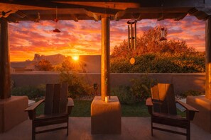 An outdoor patio with two wooden chairs, a hanging wind chime, and a view of a sunset over a mountain range.