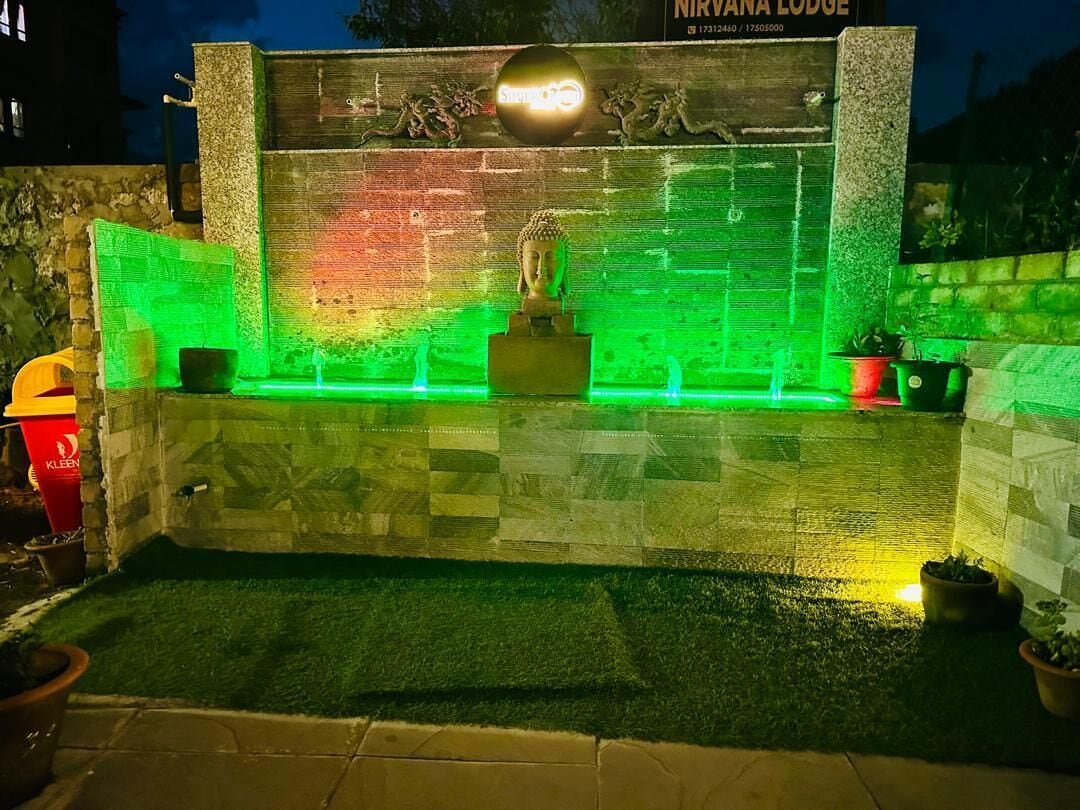 A Buddha statue in a well-lit alcove with a green-lit backdrop, surrounded by stone walls and potted plants.