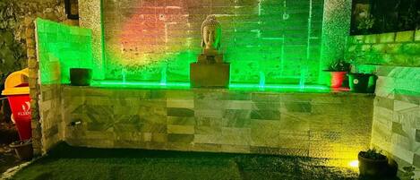 A Buddha statue in a well-lit alcove with a green-lit backdrop, surrounded by stone walls and potted plants.
