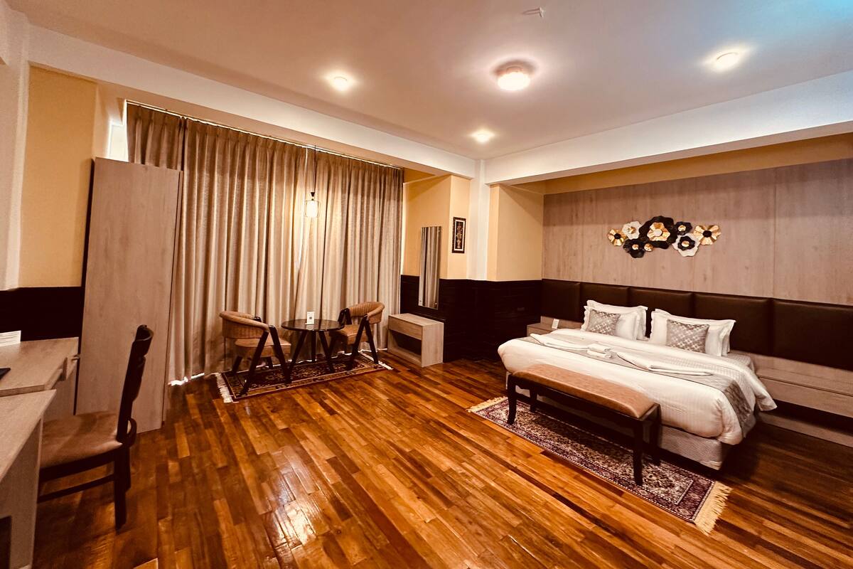 A modern bedroom with a wooden floor, a bed with white bedding, a headboard with a floral design, and a large window with curtains.