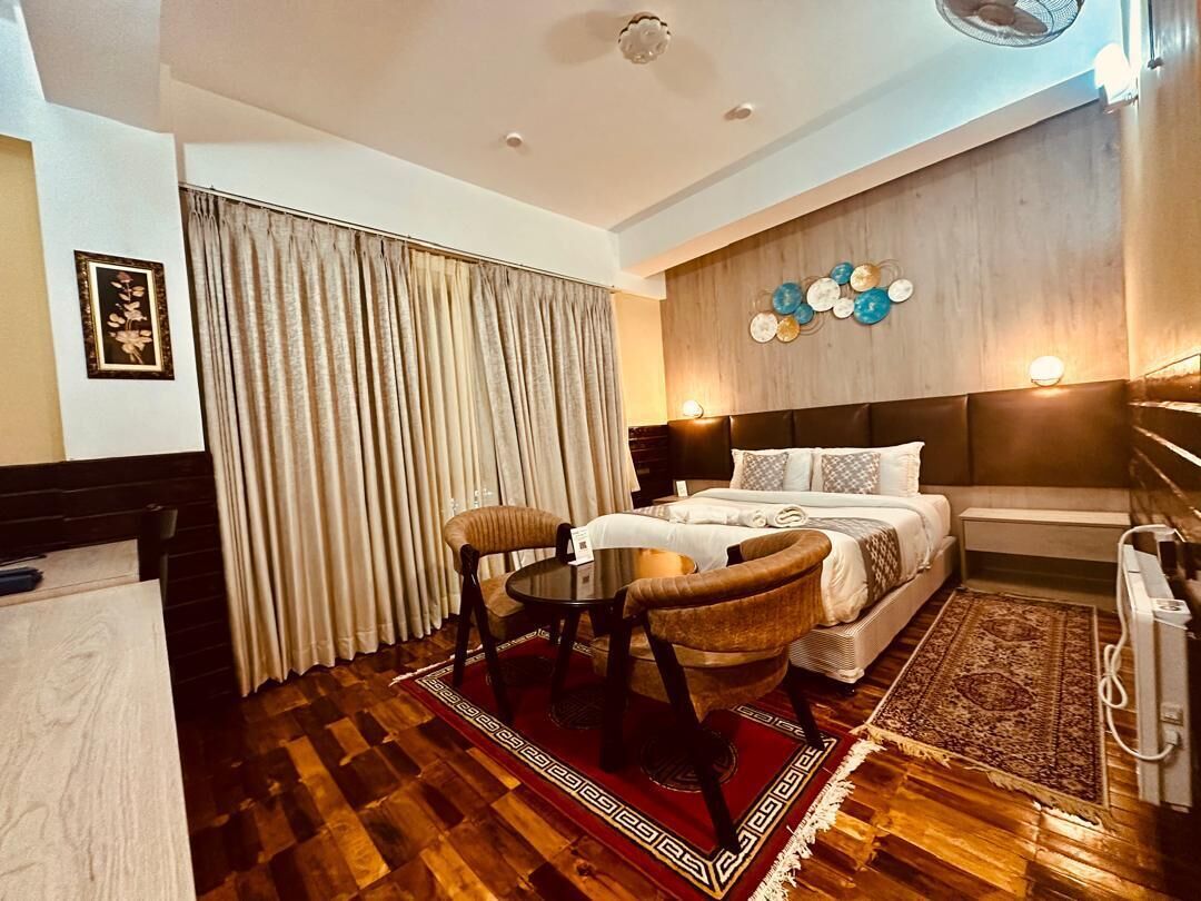 A hotel room with a bed, a desk, a chair, a small table, a rug, and a wall-mounted light fixture.