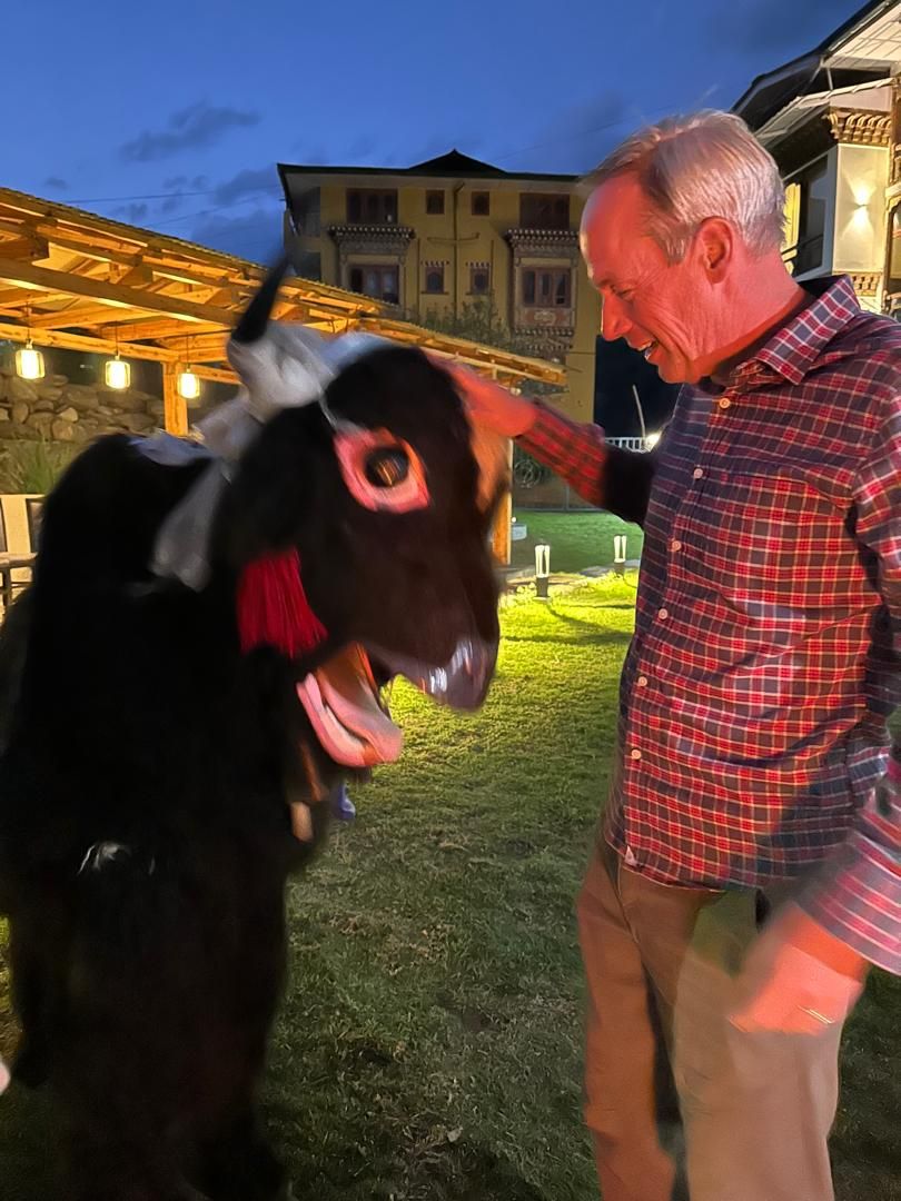 Eine Person interagiert mit einem Esel in einer offenen Landschaft, im Hintergrund ist ein Gebäude zu sehen.