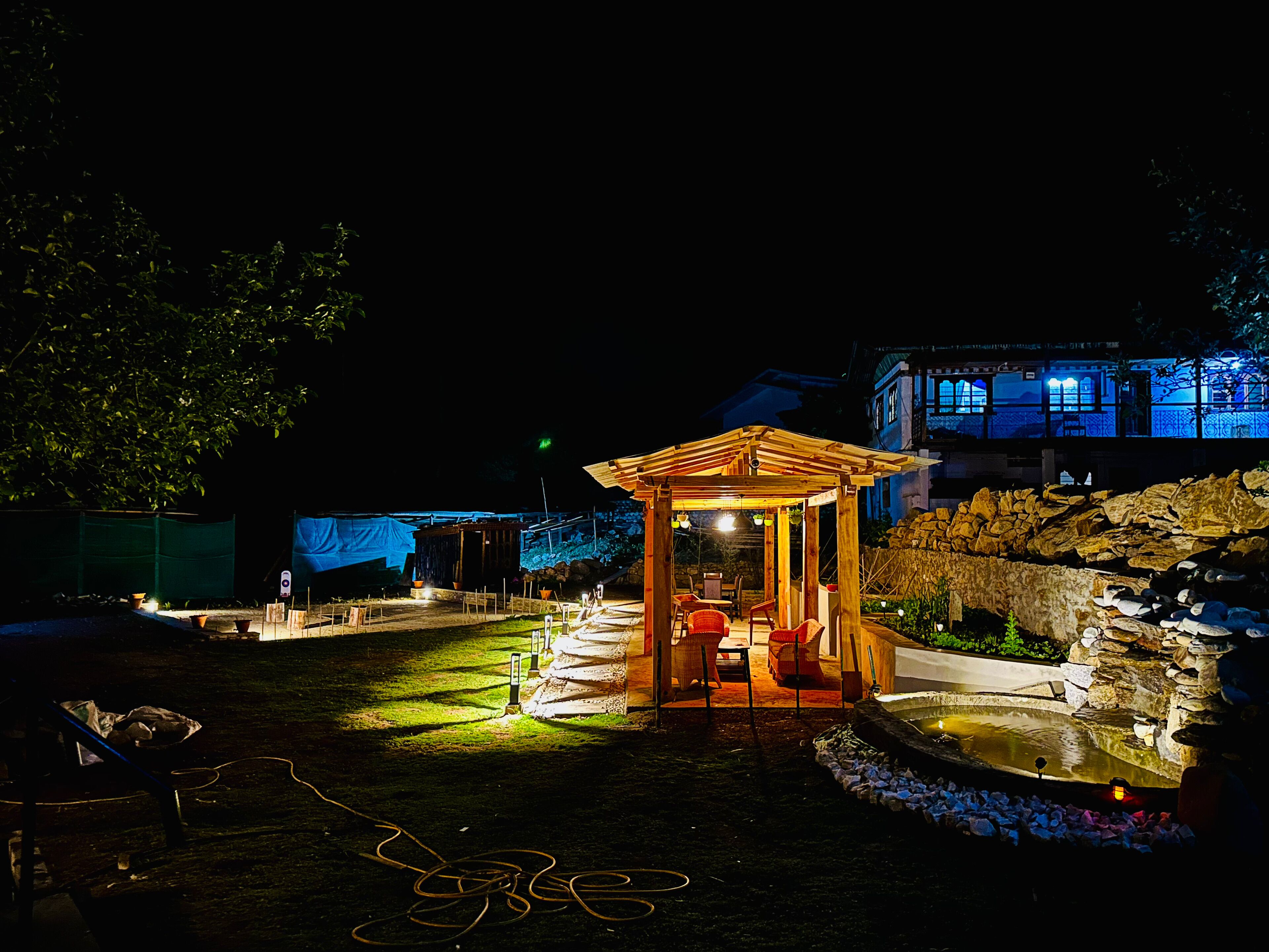 A gazebo with a wooden roof and a small table inside, surrounded by a garden with a pond and rocks.