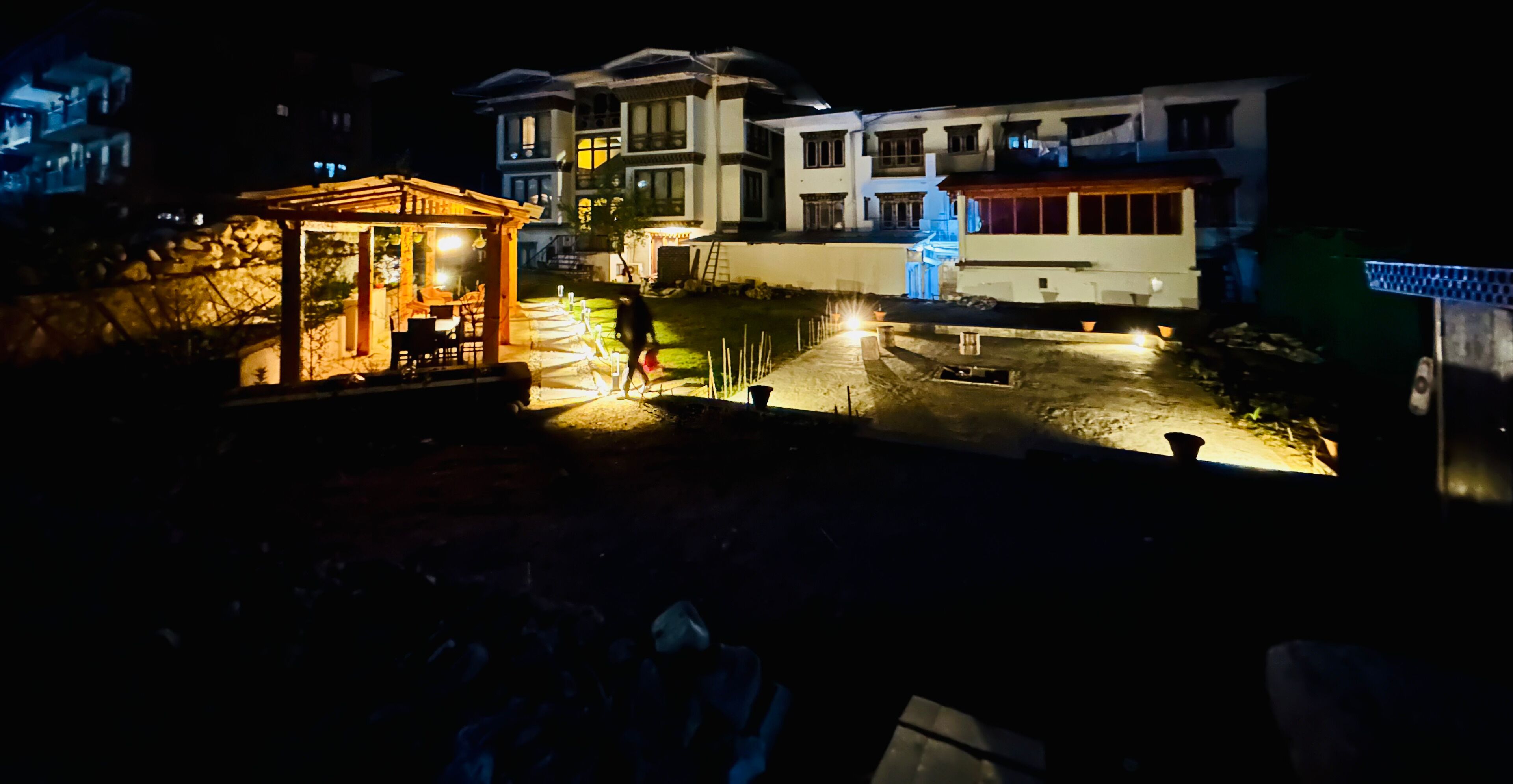 Vista nocturna de una zona residencial con un quiosco iluminado y un camino pavimentado que conduce a una casa.