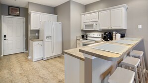 Une cuisine moderne avec des meubles blancs, un réfrigérateur, un four à micro-ondes, une cuisinière et un coin bar avec des tabourets.