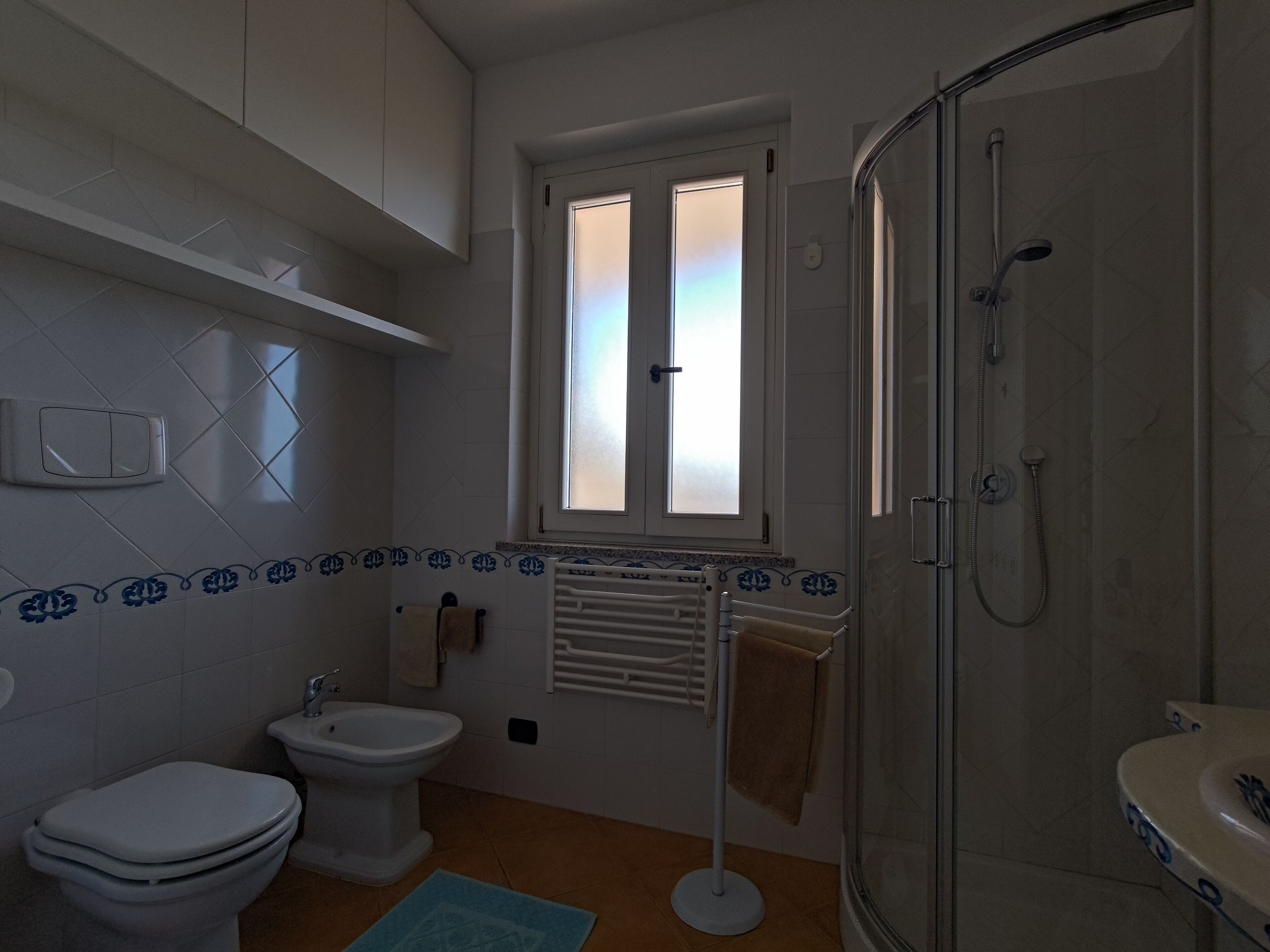 A bathroom with a toilet, bidet, and shower. There is a window with frosted glass, a towel rack, and a radiator.