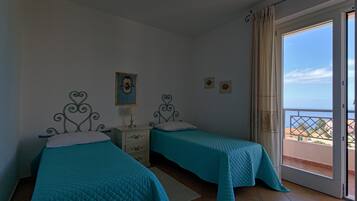 A bedroom with two beds, a nightstand, a window with a view of the sea, and a door to a balcony.