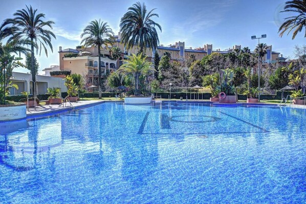 A large outdoor swimming pool surrounded by palm trees and potted plants, with buildings in the background.