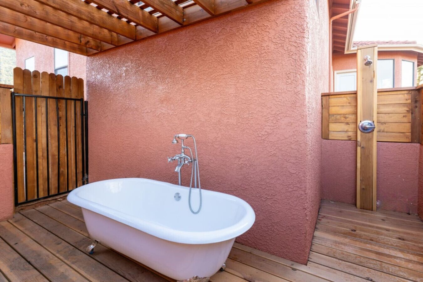 A white freestanding bathtub with a showerhead, set on a wooden deck with a pink textured wall and a wooden gate in the background.
