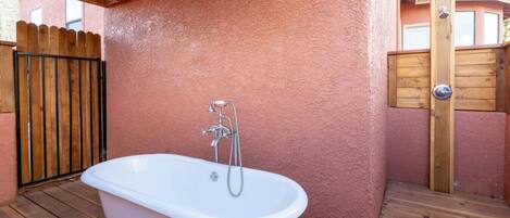 A white freestanding bathtub with a showerhead, set on a wooden deck with a pink textured wall and a wooden gate in the background.