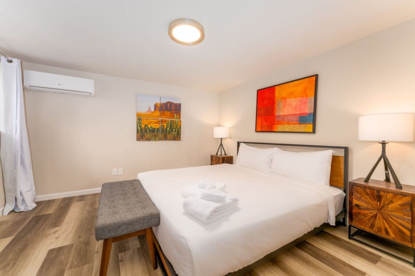 Una habitación de hotel con una cama grande, un banco, una mesita de noche, una lámpara y dos obras de arte en la pared.