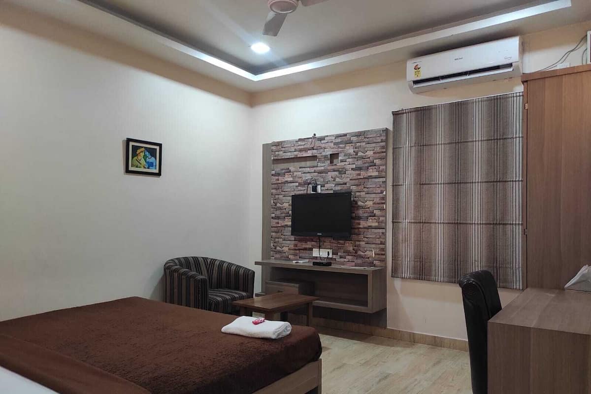 A hotel room with a bed, a TV mounted on a stone wall, a desk, a chair, and an air conditioning unit.