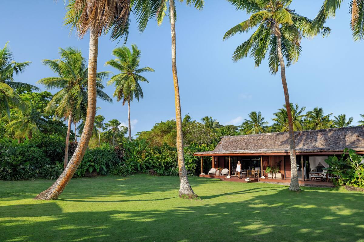 A tropical resort with a thatched-roof building, lush green lawn, and tall palm trees.