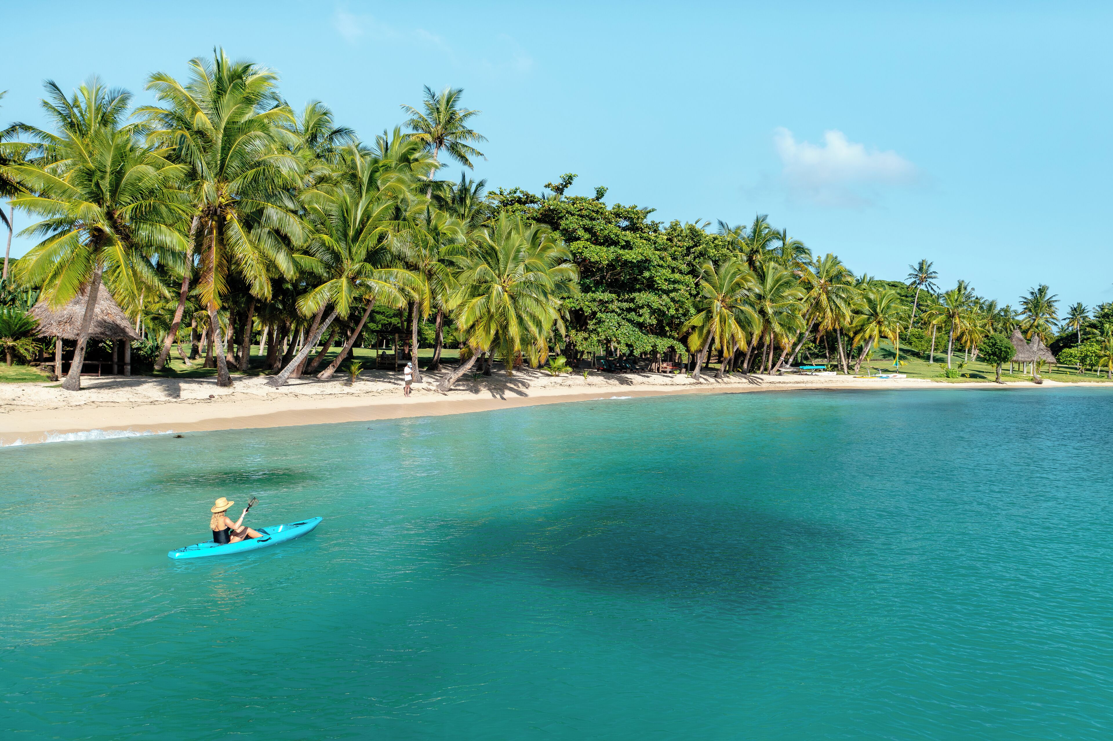 A clear turquoise sea with a person kayaking, palm trees, and a beach.