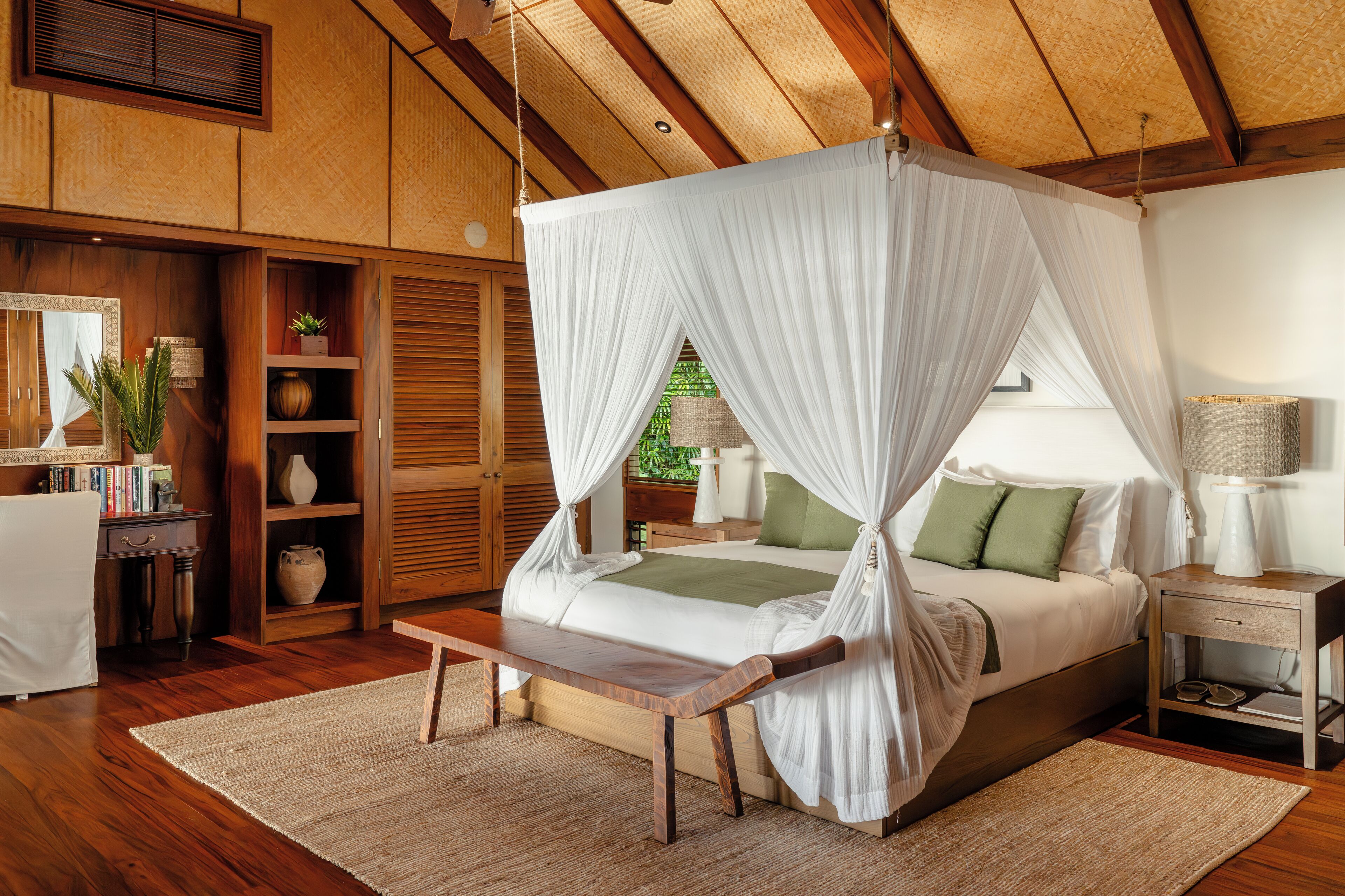 A bedroom with a canopy bed, wooden flooring, a bedside table, a nightstand, and a small window.