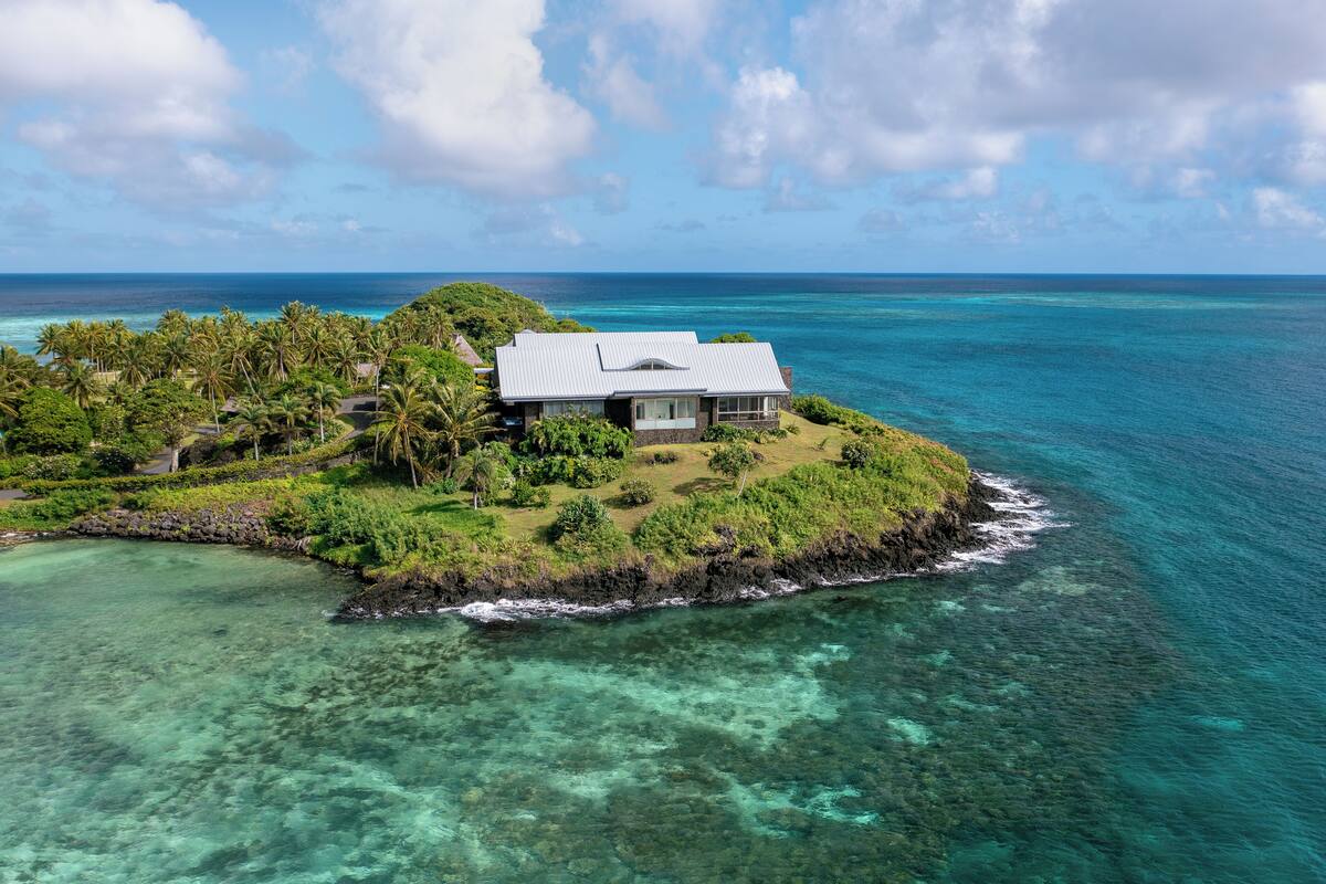 An aerial view of a tropical island with a single house surrounded by lush greenery and clear turquoise waters.