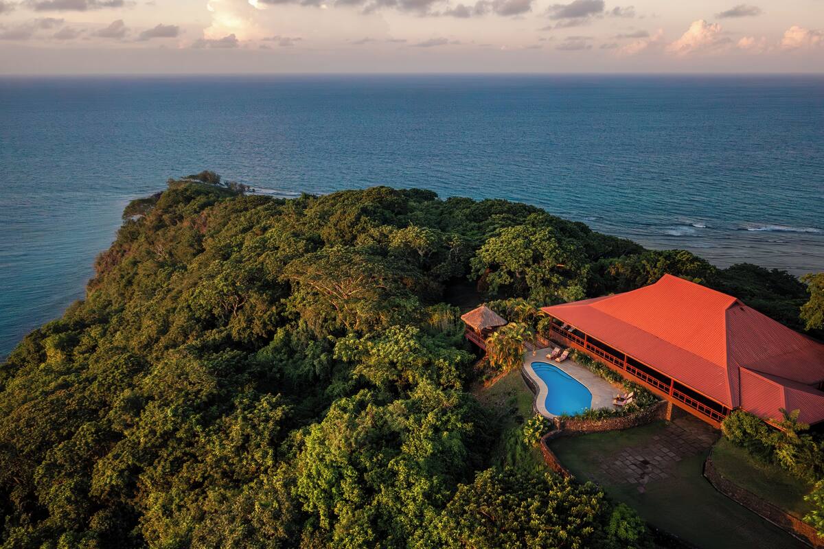 A large, red-roofed building surrounded by lush greenery, with a swimming pool and a view of the ocean.