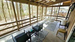 Un espace extérieur abrité, équipé d’une table et de chaises, d’un ventilateur de plafond et offrant une vue sur la forêt environnante.