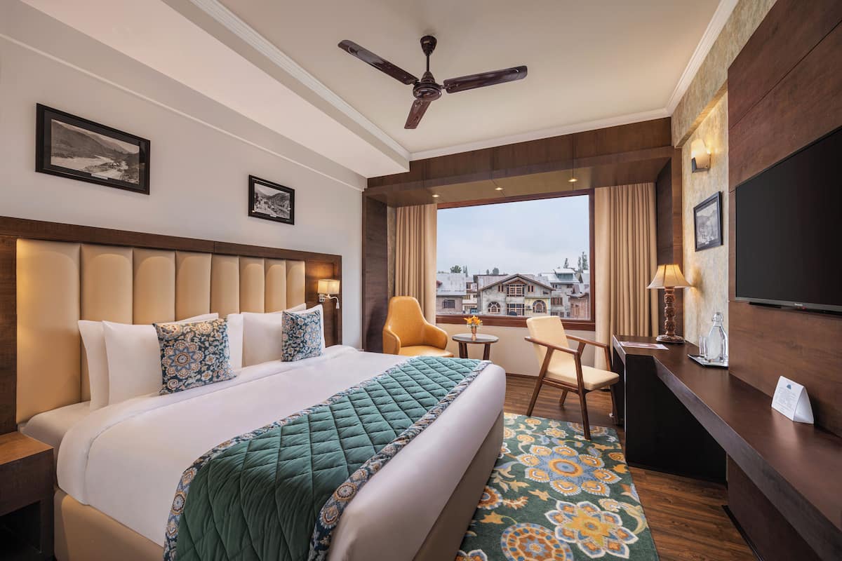 A hotel room with a large bed, a desk, a chair, a ceiling fan, and a view of buildings through the window.